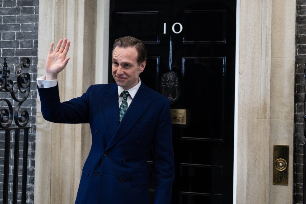 Rafe Spall as the Prime Minister standing waving and smiling outside of the door of 10 Downing Street